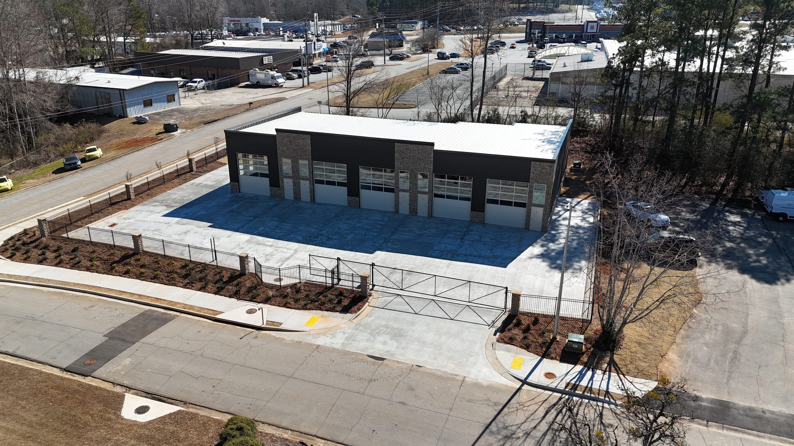 Angled aerial view of the facility, driveway gate, and surrounding commercial area